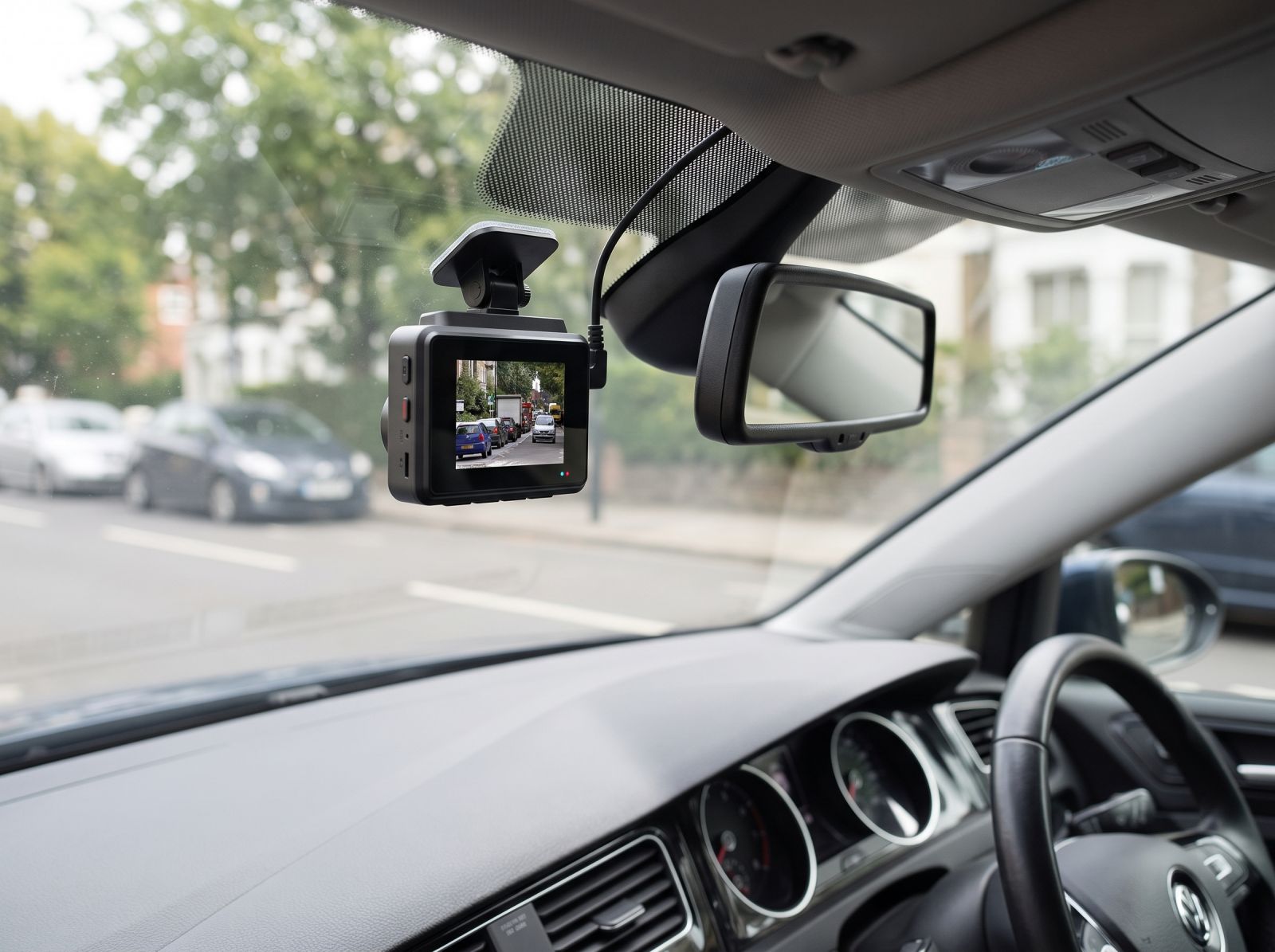 Black dash cam mounted on a car windshield beside the rearview mirror, with a built-in screen displaying recorded road footage.