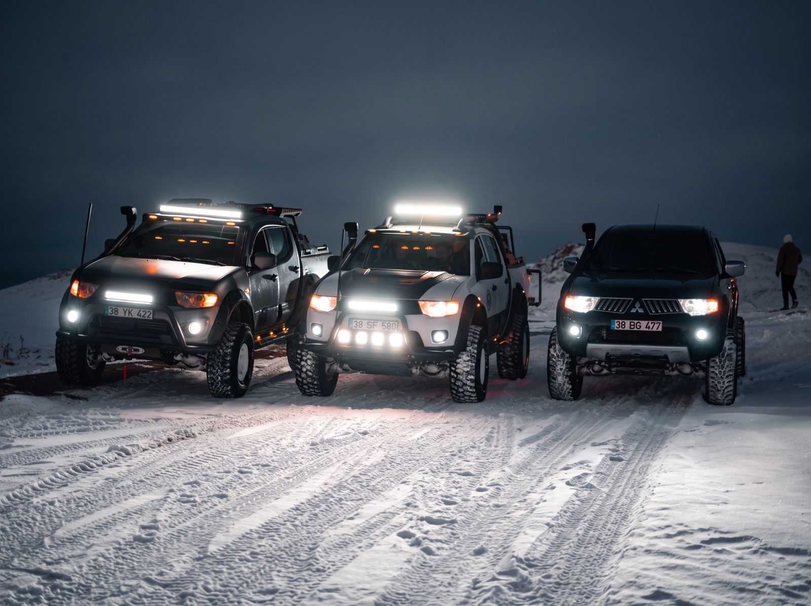Three off-road Mitsubishi 4WD vehicles equipped with bright LED light bars and driving lights parked on a snowy mountain trail at dusk.