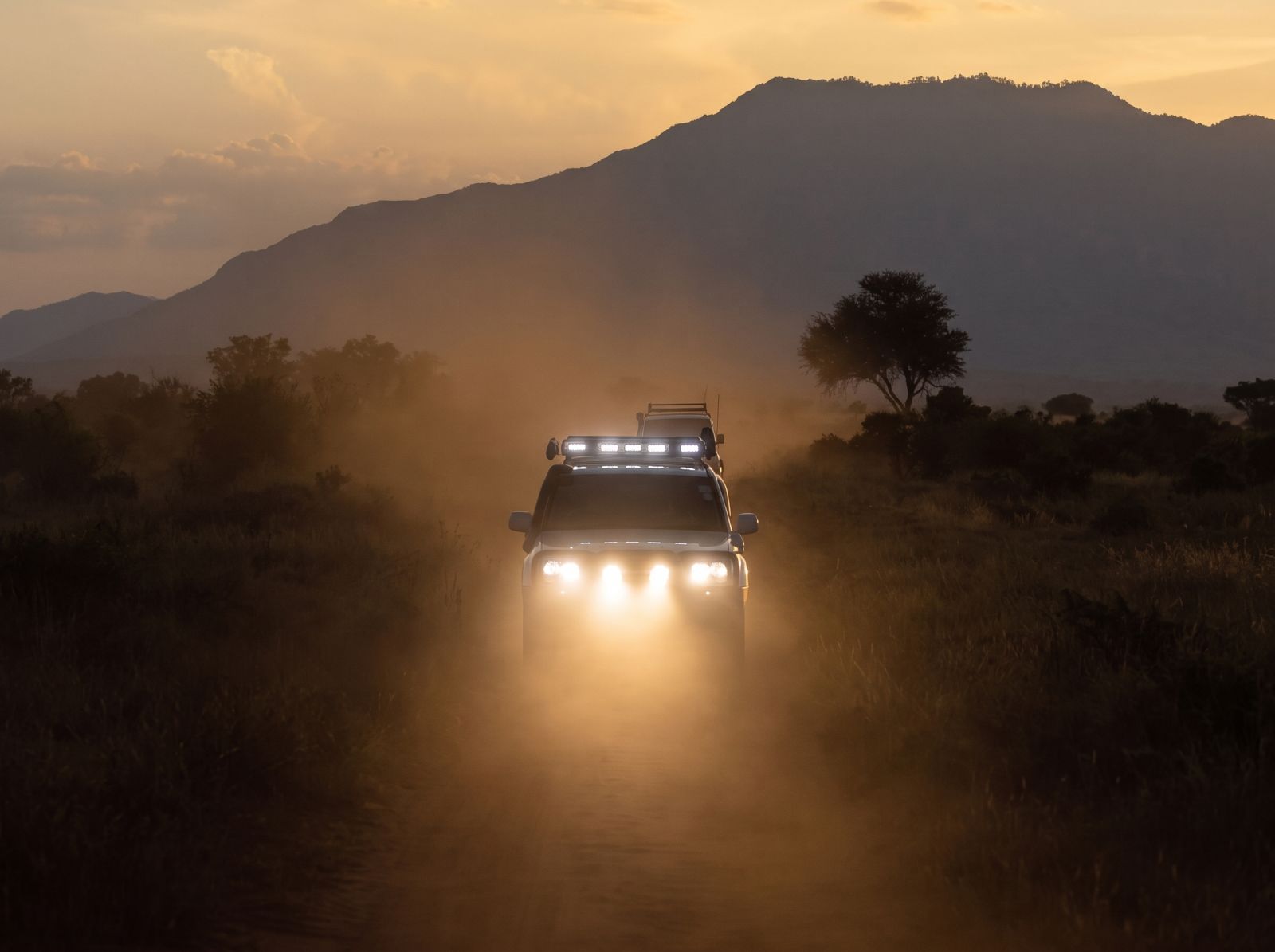 Off-road 4x4 vehicles with bright LED lights driving along a dusty trail at sunset, surrounded by mountains and rugged wilderness.